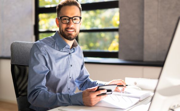 Imagem de homem de óculos segurando um celular e fazendo anotações em um fichário